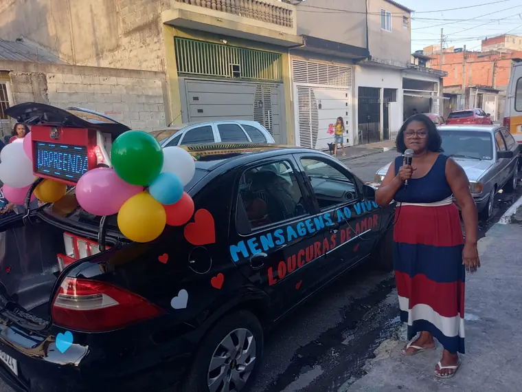Carro de som decorado com balões coloridos e faixa Mensagem ao Vivo, com a homenageada ao lado